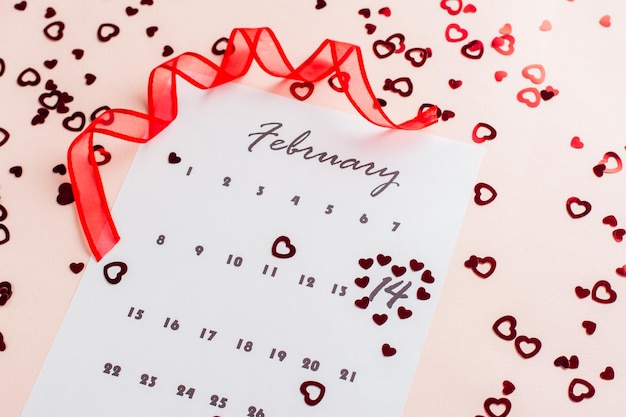 Día de San Valentín. Los pequeños corazones rojos resaltan la fecha 14 de febrero en una hoja de calendario y una cinta roja sobre un fondo rosa lleno de corazones. De cerca