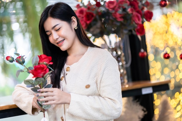 Día de San Valentín Retrato Feliz Sonriente hermosa joven asiática con ropa abrigada sosteniendo un ramo de rosas rojas con un jarrón de rosas en la sala de estar o en la cafetería