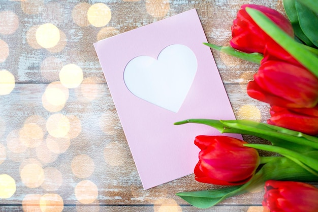 día de san valentín, saludo, amor y concepto de vacaciones - cierre de tulipanes rojos y tarjeta de saludo con corazón sobre luces