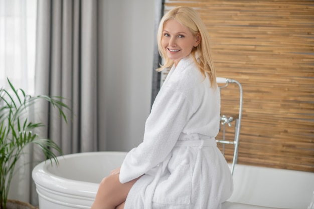 Día de spa. Mujer bonita rubia en una bata de baño blanca preparando el baño