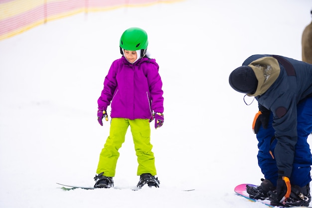 En el día del viento frío en la estación de esquí de montaña, el padre enseña a su hija pequeña a hacer snowboard