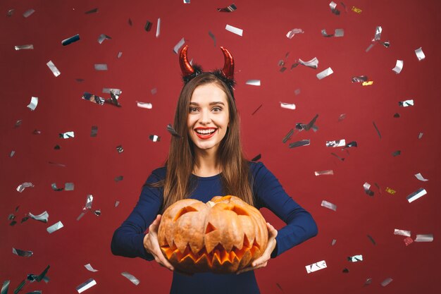 Diablo de Halloween con una calabaza tallada - aislada sobre fondo rojo. Mujer joven emocional en disfraces de Halloween.