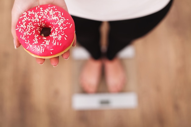 Diät. Frauen-messendes Körpergewicht auf der Waage, die Donut hält.