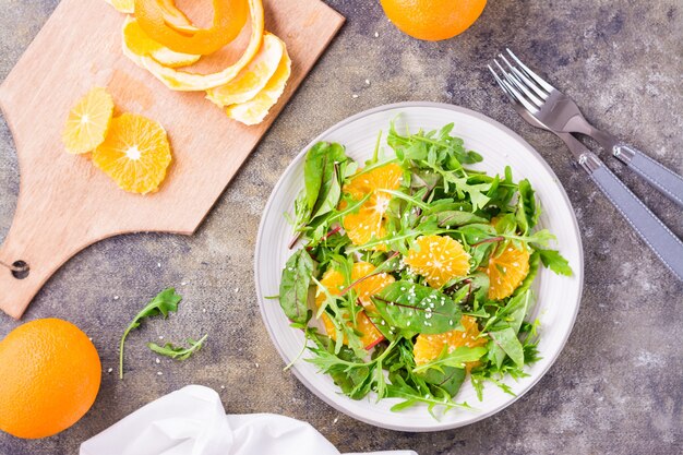 Diät-vegetarischer Vitaminsalat aus Orangenscheiben und einer Mischung aus Rucola, Mangold und Mizun-Blättern auf einem Teller und einem Schneidebrett mit geschälter Orange auf dem Tisch. Ansicht von oben