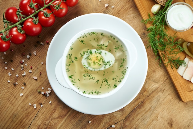 Diätetische Suppe mit Huhn, gekochtem Eiergemüse und Petersilie in Kompostion mit Zutaten auf Holzoberfläche in weißer Schüssel. Draufsicht leckere Suppe. Flaches Essen