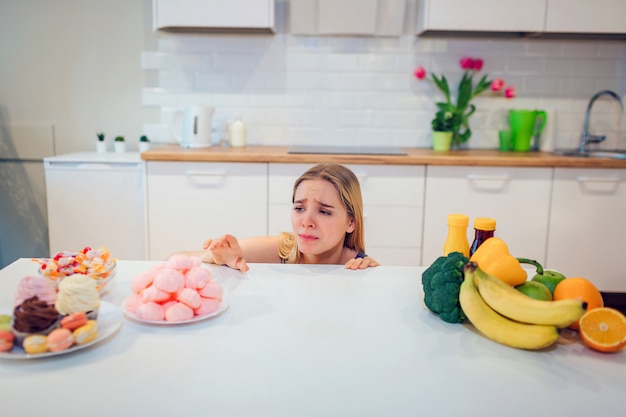 Diätkampf. Junge traurige Frau im blauen T-Shirt, das zwischen frischem Obstgemüse oder Süßigkeiten in der Küche wählt. Wahl zwischen gesundem und ungesundem Essen. Diät halten. Gesundes Essen