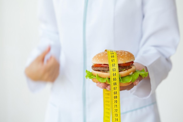 Diätkonzept, Doktor, der schädlichen fetten Burger mit gelbem Maßband hält