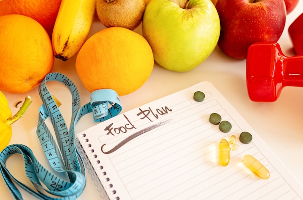 Diätplan, Menü oder Programm, Gewichtsverlust, Maßband, Hanteln und diätetische Lebensmittel frisches Obst auf einem weißen Tisch
