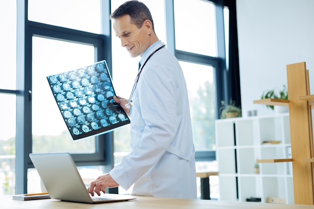 Diagnósticos médicos. Radiologista sério e inteligente segurando uma radiografia e pressionando um botão em seu laptop enquanto está no escritório