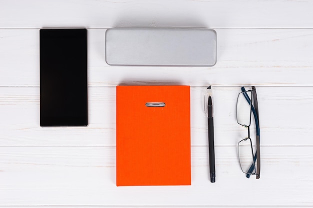 Diario naranja con un bolígrafo para concertar citas, organizar un horario, gafas elegantes y un estuche para gafas cerca del teléfono móvil en la mesa de madera blanca