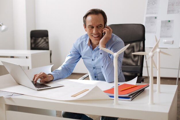 Dias de workign positivos. Engenheiro alegre e encantado sentado à mesa e falando no celular enquanto trabalhava no laptop