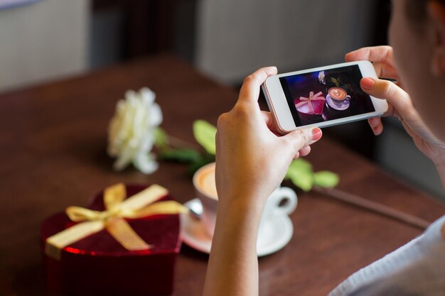 días festivos, día de san valentín, tecnología y concepto de personas: cerrar las manos con un teléfono inteligente tomando una caja de regalo en forma de corazón y una foto de una taza de café