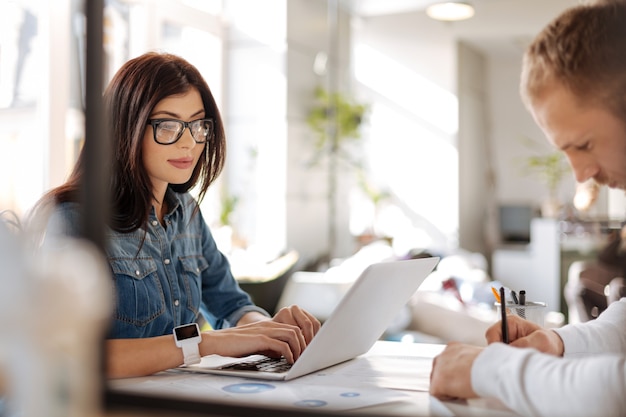 Días laborales habituales. Mujer hermosa trabajadora seria sentada en la mesa y escribiendo en la computadora portátil mientras trabaja en la oficina