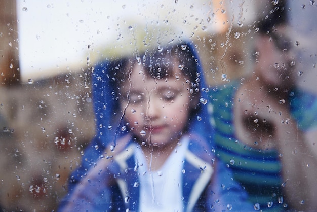 Días lluviosos sin nada que hacer Un niño mirando hacia abajo detrás de una ventana con rayas de lluvia mientras su madre se sienta en el fondo