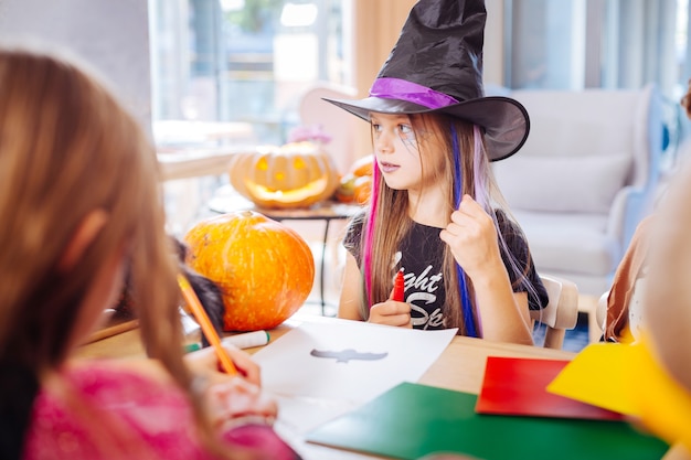 Dibujo de murciélago. Hermosa chica de cabello oscuro con labios morados con sombrero de mago negro para Halloween dibujando murciélago negro