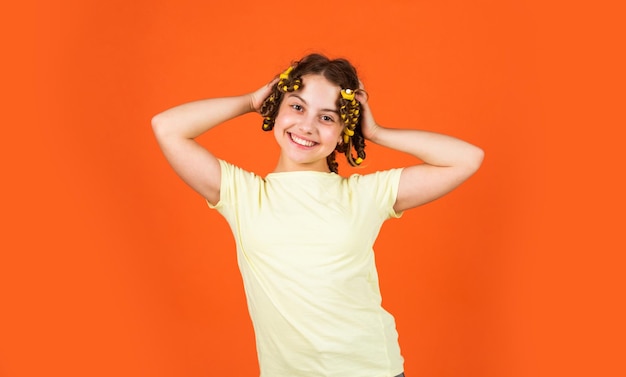 Dicas de estilo Menina pequena ondulando o cabelo usando rolinhos fundo laranja Filha com rolinhos na cabeça rindo Salão de cabeleireiro Rotina de beleza feminina Penteado de criança adorável Hobbies adolescentes