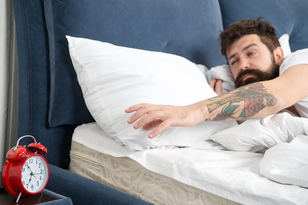 Dicas para acordar cedo Homem barbudo hipster cara sonolento cama com despertador Desligue esse toque Que barulho terrível Problema acordar cedo Acorde com despertador Dormi demais de novo