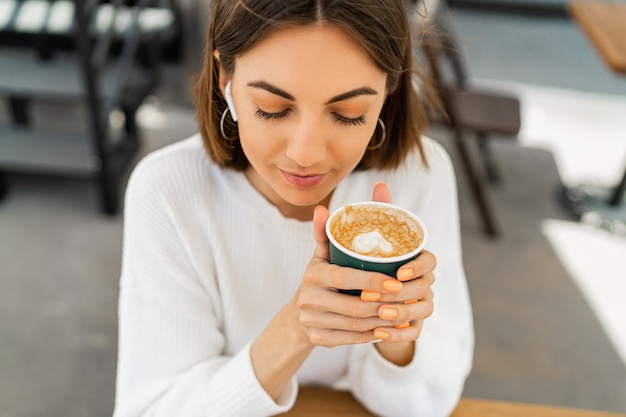 Dichosa mujer de pelo corto disfrutando de un capuchino en el café, vistiendo un acogedor suéter blanco