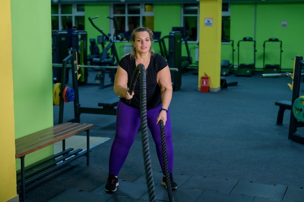 Dicke Frau beim Krafttraining mit Kampfseilen im Fitnessstudio. Ein fettleibiger Athlet bewegt die Seile in einer Wellenbewegung, um die Fettverbrennung zu trainieren