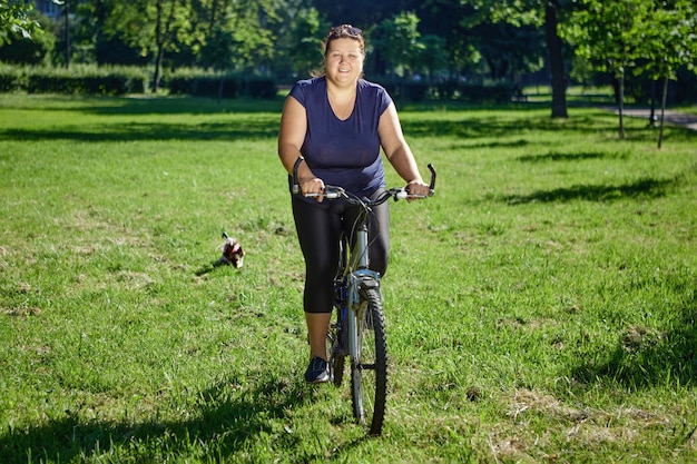 Dicke weiße Frau, die im öffentlichen Park des sonnigen Sommertages radfährt