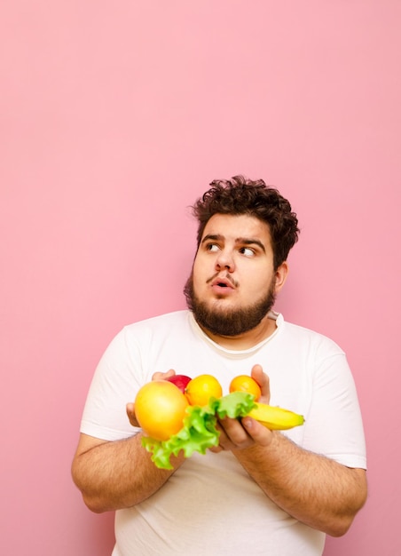 dicker mann auf diät, der mit früchten in den händen auf rosa hintergrund steht und auf kopienraum schaut