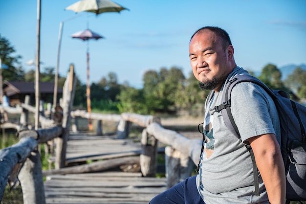 Dicker Mann des asiatischen Rucksacktouristen mit Bart auf der Holzbrücke mit blauem Himmel in der Ferienzeit