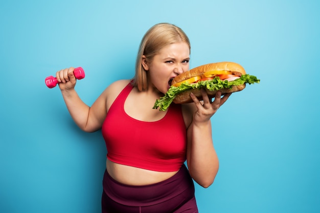 Dickes Mädchen macht Fitnessstudio und will ein Sandwich essen. Konzept der Unentschlossenheit und des Zweifels