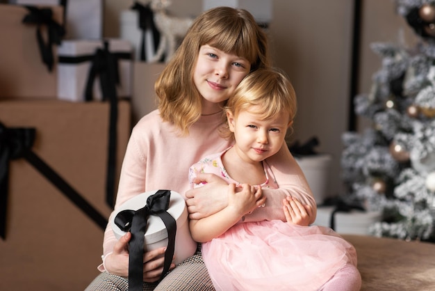 Die ältere Schwester umarmt die jüngere vor dem Hintergrund eines Weihnachtsbaums und eines Haufens von Geschenken