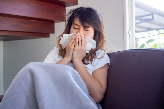 Die asiatische Frau saß krank auf dem Sofa im Haus und hatte Fieber, Husten, Niesen und eine verstopfte Nase. Bedecken Sie die Nase mit einem Stück Papier.