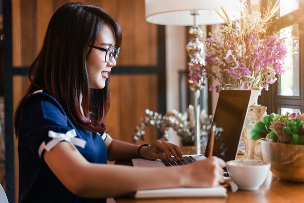 Die asiatische Geschäftsfrau, die mit Laptop arbeitet, machen eine Anmerkung in der Kaffeestube wie dem Hintergrund.