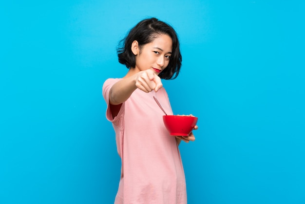 Die asiatische junge Frau, die eine Schüssel Getreide hält, zeigt Finger auf Sie mit einem überzeugten Ausdruck