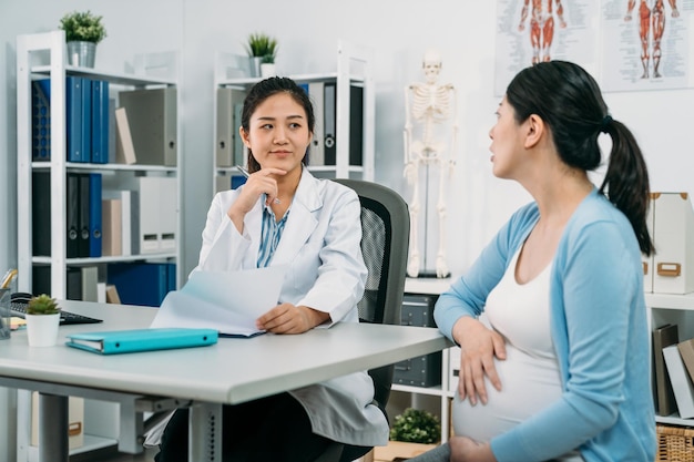 Die asiatische Ärztin reibt sich das Kinn und lächelt selbstbewusst, während sie ihrer erwartungsvollen Patientin während der Beratung am Schreibtisch im Klinikbüro zuhört