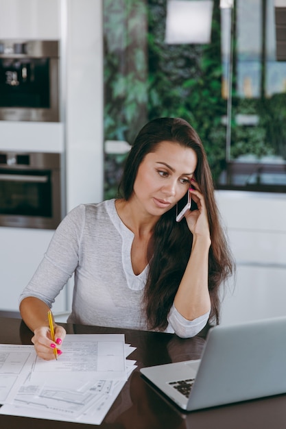 Die attraktive junge moderne Geschäftsfrau, die mit dem Handy spricht und mit Dokumenten und Laptop in der Küche zu Hause arbeitet