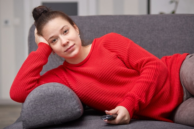 Die Aufnahme einer schwangeren Frau sieht zu Hause fern, hält die Fernbedienung und wechselt die Kanäle. Sie genießt eine interessante TV-Show und liegt auf einem grauen Sofa