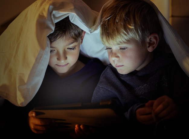 Die Augen von Kindern sehen alles frisch und voller Staunen Aufnahme von zwei entzückenden kleinen Jungen, die nachts in ihrem Schlafzimmer ein digitales Tablet unter einer Decke benutzen