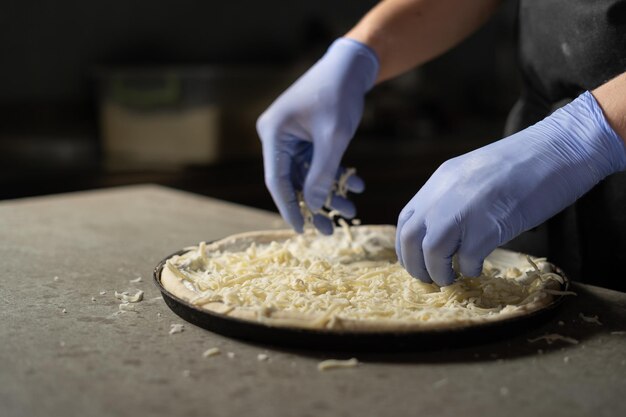 Die behandschuhten Hände des Küchenchefs legen geriebenen Käse auf eine runde vegetarische Pizza. Zutaten in einer Pizzeria. Italienisches Essenskonzept.