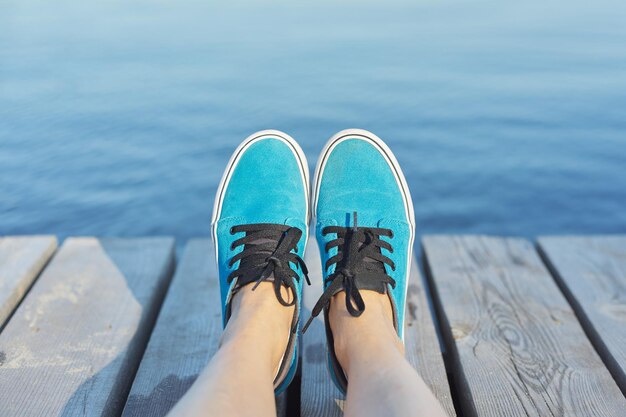 Die Beine der Frau in den blauen Turnschuhen auf hölzernem Pier, Flusssee-Teichhintergrund für Kopienraum. Urlaub, Natur, Lifestyle, People-Konzept