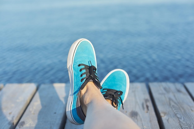 Die Beine der Frau in den blauen Turnschuhen auf hölzernem Pier, Flusssee-Teichhintergrund für Kopienraum. Urlaub, Natur, Lifestyle, People-Konzept