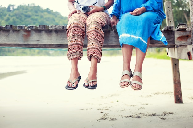 Die Beine der Hippie-Frauen sitzen auf hölzerner Brücke auf dem schönen Strand