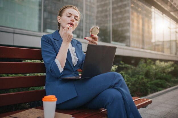 Die beschäftigte Frau hat es eilig, sie hat keine Zeit, sie wird sich schminken und am Laptop arbeiten. Arbeiter, der auf der Straße Lippenstift aufträgt und gleichzeitig am PC arbeitet.