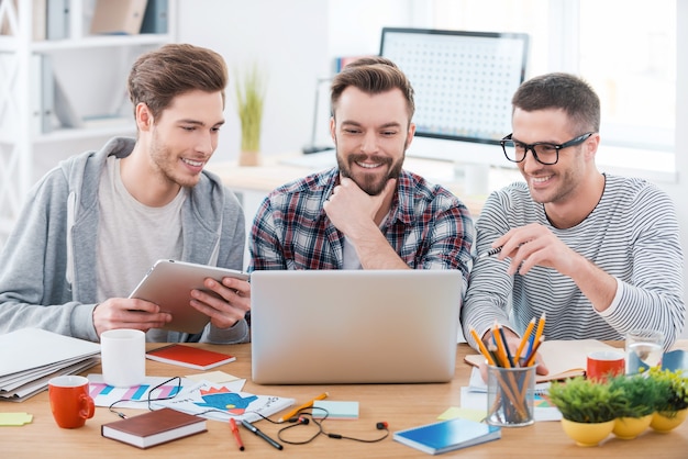 Die besten Entscheidungen mit guter Laune. Drei junge glückliche Männer, die zusammenarbeiten, während sie an ihrem Arbeitsplatz im Büro sitzen sitting