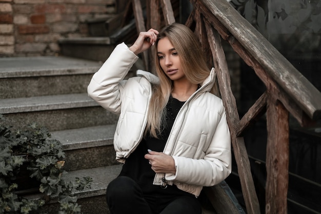 Foto die blonde frau in einer weißen jacke in schwarzen, eleganten jeans ruht auf einer vintage-steintreppe