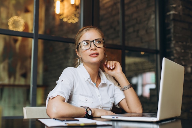 Die blonde Geschäftsfrau, die auf Arbeitsplatz sitzt und Laptop im Büro verwendet, betrachtet sie einen Ordner mit Dokumenten, spricht telefonisch und trinkt Kaffee.