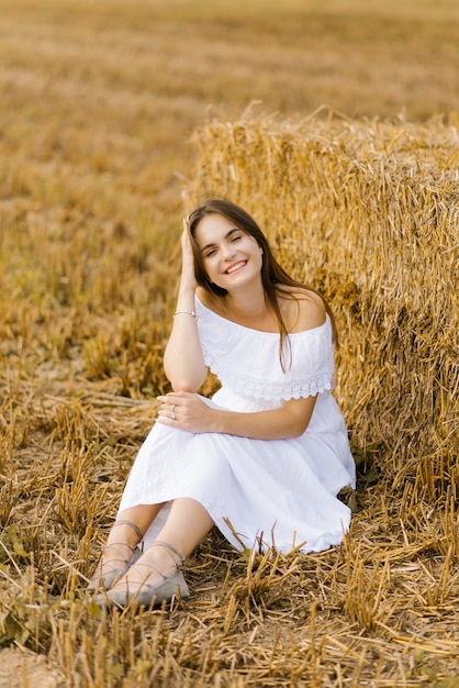 Die blonde junge Frau in einem weißen Kleid auf einem Feld mit Strohhaufen genießt einen Sommertag und lächelt