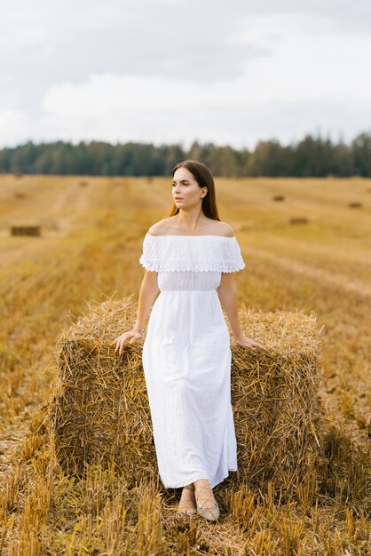 Die blonde junge Frau in einem weißen Kleid auf einem Feld mit Strohstapeln genießt einen Sommertag