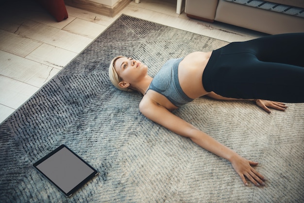 Die blonde sportliche Frau, die Sportbekleidung trägt, macht Fitnessübungen auf dem Boden unter Verwendung einer Tablette