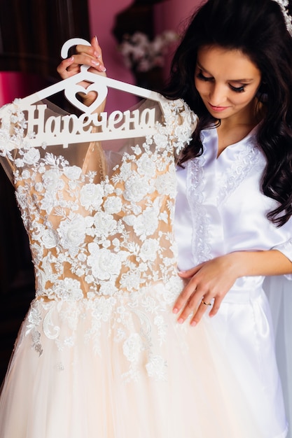 Die Braut hält einen Kleiderbügel mit einem Hochzeitskleid und schaut es sich an.