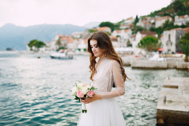 Die Braut hält einen Rosenstrauß in den Händen und steht auf dem Pier in der Nähe der Altstadt von Perast