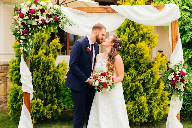 Die Braut in einem weißen Kleid mit Spitze hält einen Hochzeitsstrauß und der Bräutigam sieht sie an und möchte sie küssen und hinter ihnen ist ein Holzbogen mit Ornamenten