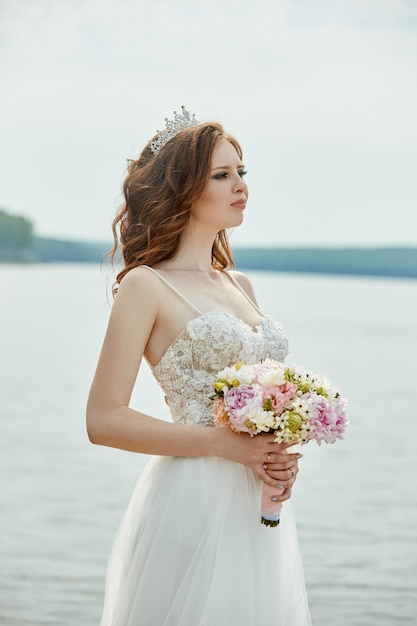 Die Braut in einem weißen Kleid steht auf der Brücke und schaut auf den See. Frau in einem Hochzeitskleid mit einem Blumenstrauß